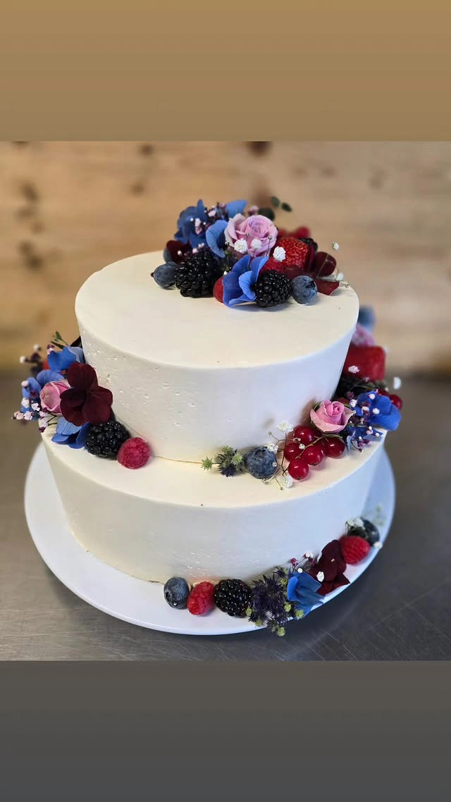 Zweistöckige Hochzeitstorte in Weiß mit Beeren, Rosen sowie blauen und burgunderfarbenen Akzenten