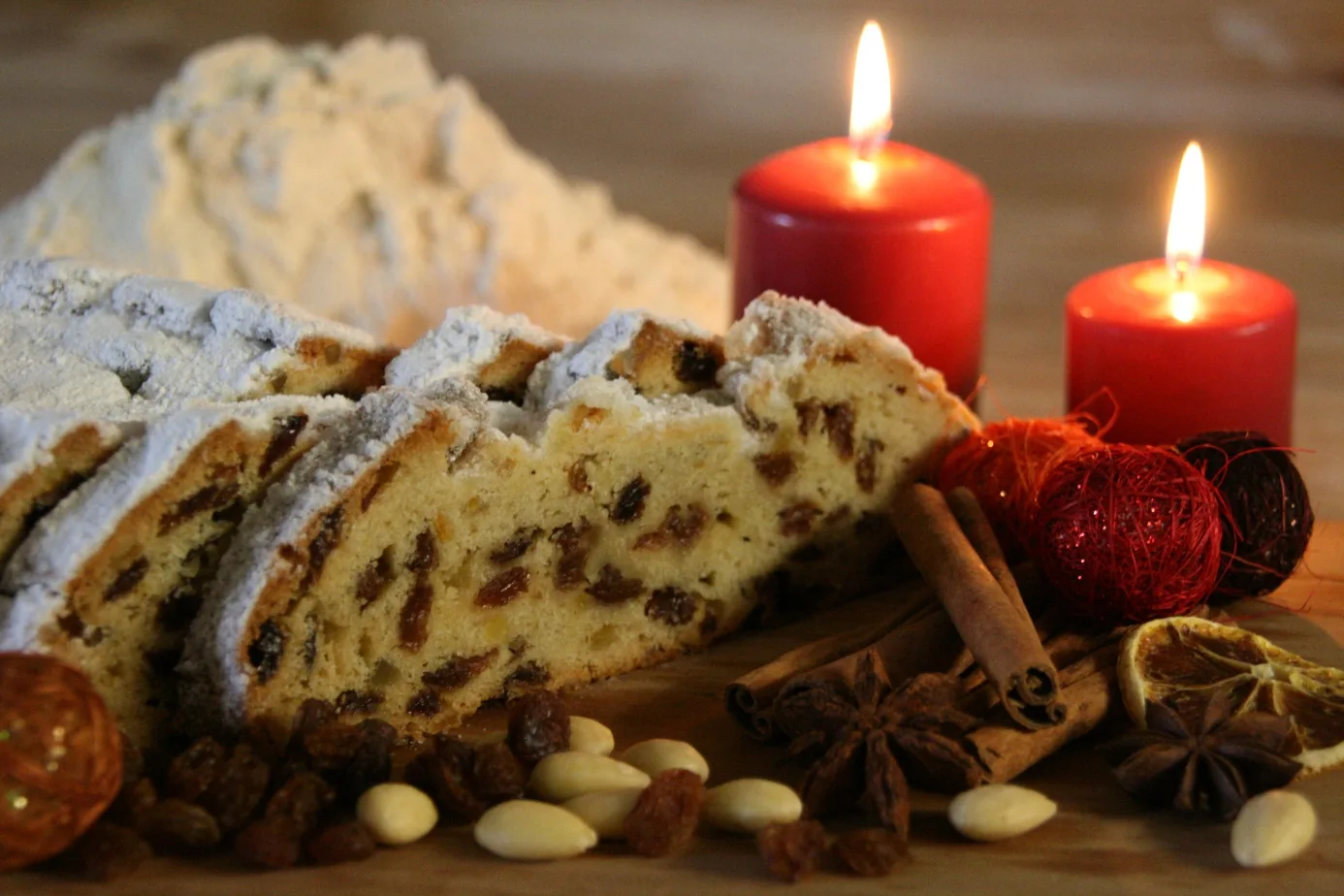 Angeschnittener Dresdner Christstollen der Landbäckerei Friedrich mit Rosinen, daneben Mandeln, Zimtstangen und brennende Kerzen