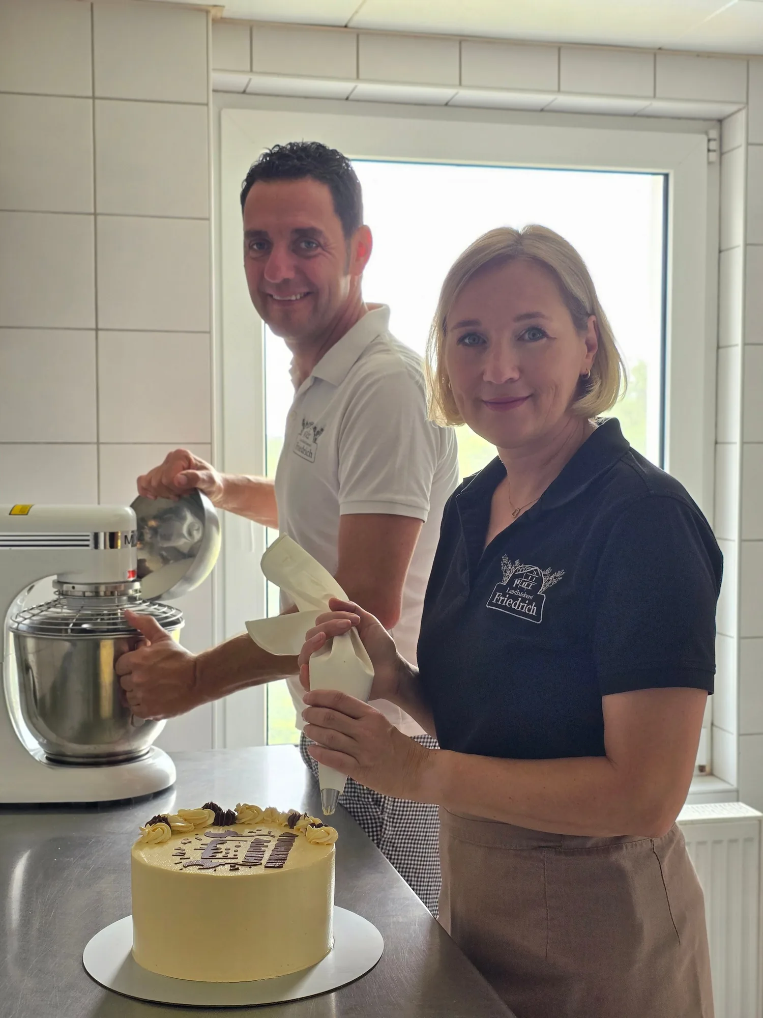 Anja Friedrich verziert eine Torte mit dem Spritzbeutel, Bäckermeister Matthias Friedrich arbeitet am Rührgerät in der Backstube der Landbäckerei Friedrich